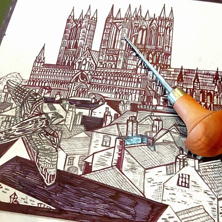 Picture shows a design of Lincoln Cathedral in black ink on lino with a peregrine falcon flying over the rooftops towards the cathedral by Printmaker Dehlia Barnard-Edmunds. There is a linocut carving tool on top of the linocut plate.
