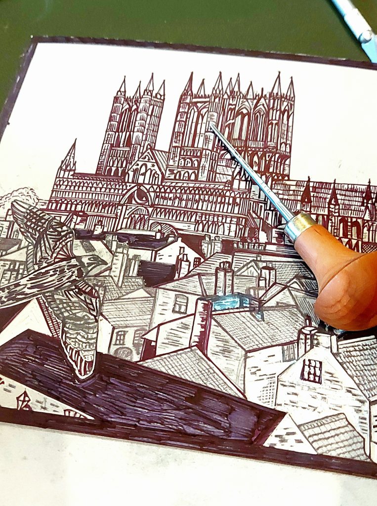 Picture shows a design of Lincoln Cathedral in black ink on lino with a peregrine falcon flying over the rooftops towards the cathedral by Printmaker Dehlia Barnard-Edmunds. There is a linocut carving tool on top of the linocut plate.