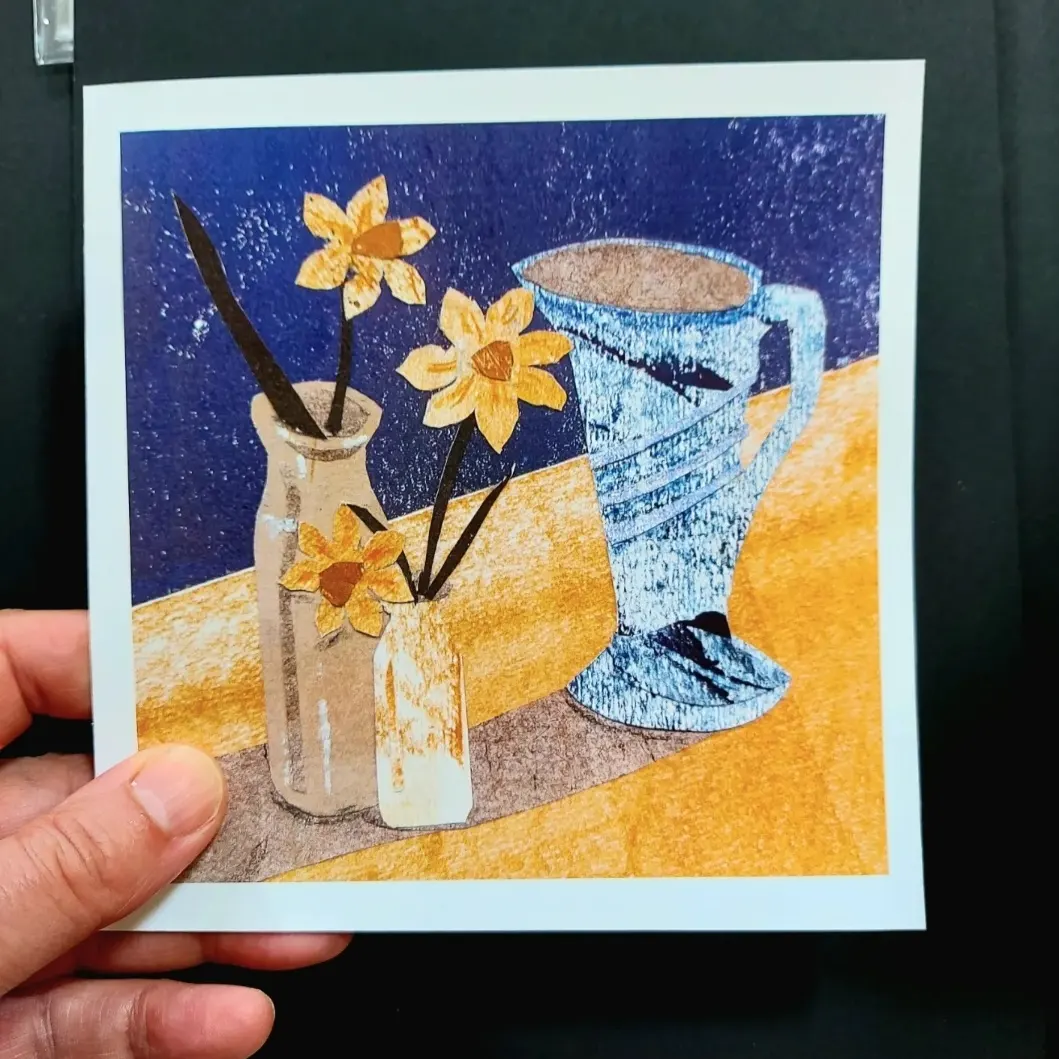 Hand holding a greetings card of a still life collage with a vintage jug and two Victorian ink pots with daffodils by Dehlia BArnard-Edmunds AKA The Merryweather Artist