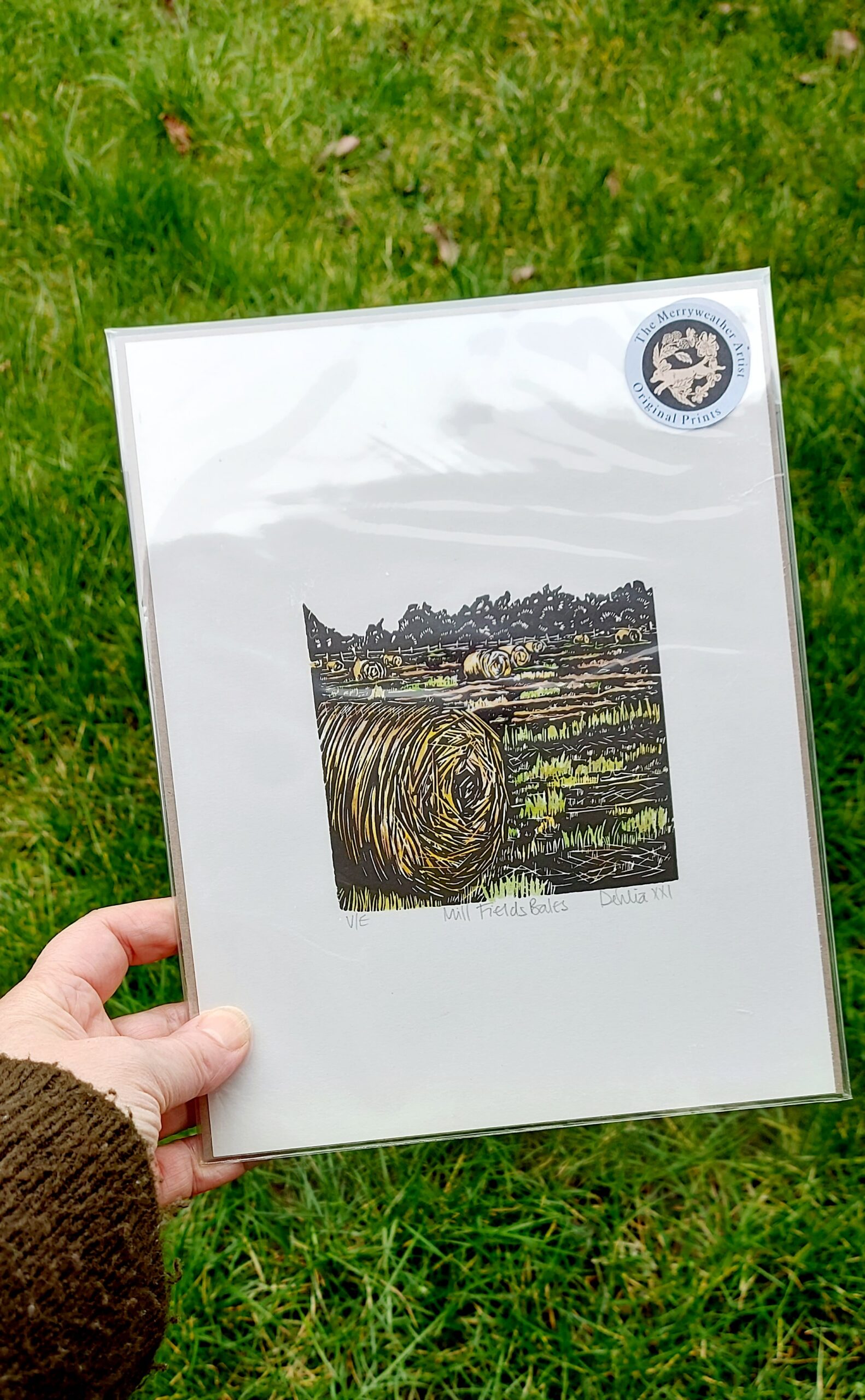 A hand holding a packaged lino print of Hay Bales by Dehlia Barnard-Edmunds AKA The Merryweather Artist on a grassy background