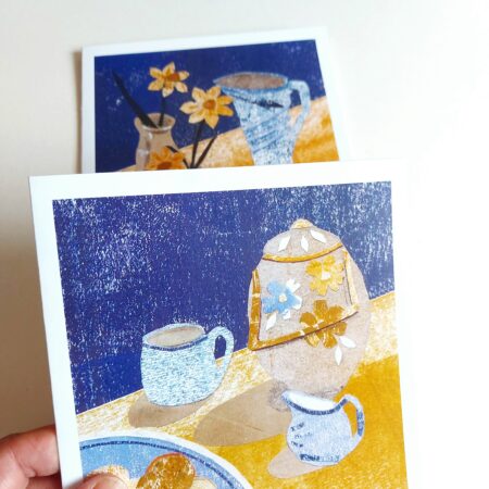 Hand holding a greetings card with collaged still life of biscuits and tea with another card in the background by Dehlia Barnard-Edmunds AKA The Merryweather Artist
