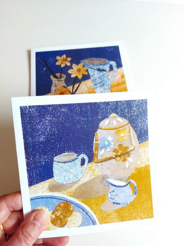 Hand holding a greetings card with collaged still life of biscuits and tea with another card in the background by Dehlia Barnard-Edmunds AKA The Merryweather Artist