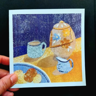 hand holding a greetings card with a blue and yellow image of a cup, a jug, a vintage biscuit barrel and a plate of biscuits collage by Dehlia Barnard-Edmunds