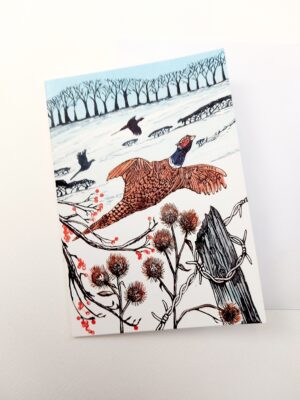 A greetings card showing an original linocut print of a winter pheasant flying across a field of snow towards a row of trees on the hill by Dehlia Barnard-Edmunds